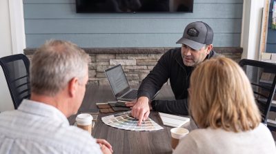A Kinmount team member in a Kinmount Exteriors cap points to siding color swatches while a couple looks on during a Lakeville exterior estimate in the showroom.